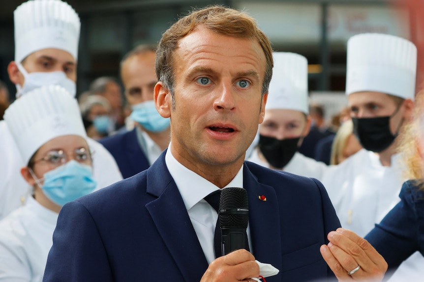 French President Emmanuel Macron wears a blue suit and holds a microphone.